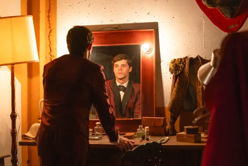 A young man in a burgundy suit studies his reflection in a dressing-room mirror, an ornate jacket and cape hanging behind him, in A Day Like No Other