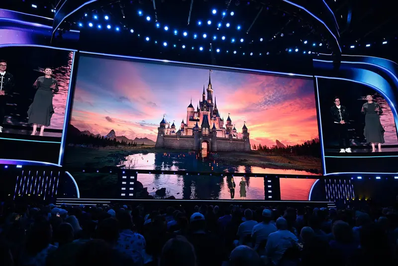 Two presenters on stage at Disney's D23 Entertainment Showcase in front of a large screen showing a Disney castle at sunset, with the audience seated below