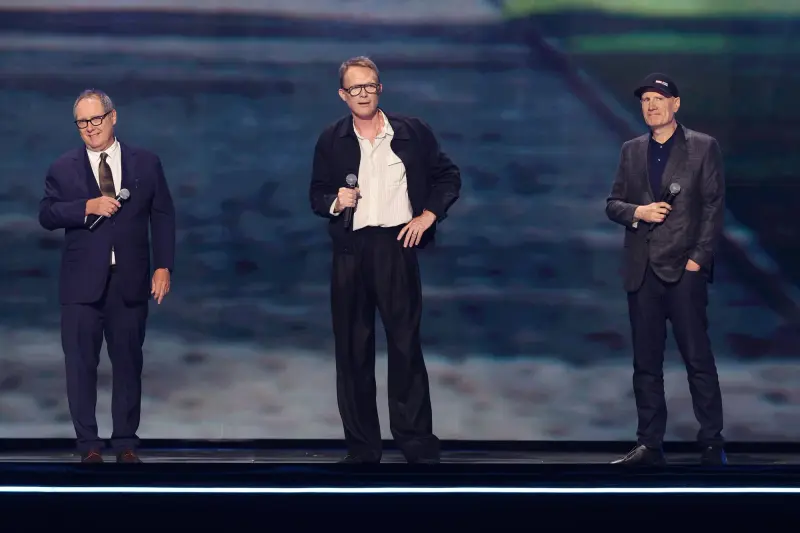 Three men holding microphones on a darkened stage at D23, with a large screen lit behind them