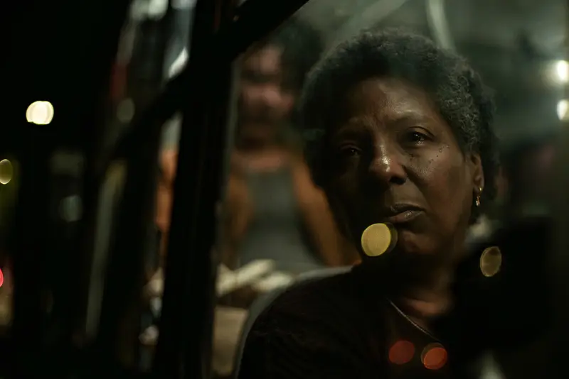 Rejane Faria as Vicentina looks through a bus window at night, city lights blurred behind her, in On Behalf of My Son