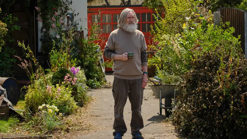 A bearded man holds a mug in an overgrown garden in Small Prophets
