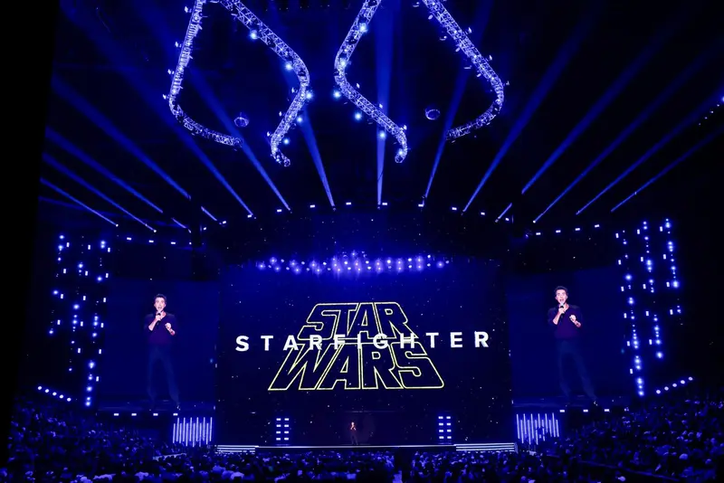 The Star Wars: Starfighter logo on a large screen at D23, lit in blue, with two speakers standing on the stage in front of a full auditorium